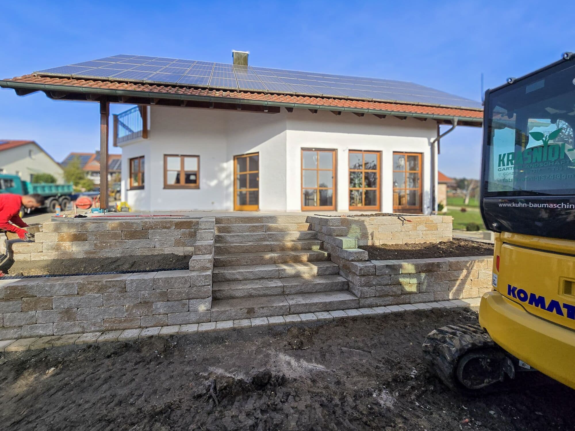 Natursteinmauer mit Treppe vor einem Wohnhaus, rechts der eigene Bagger mit Krasniqi-Beschriftung